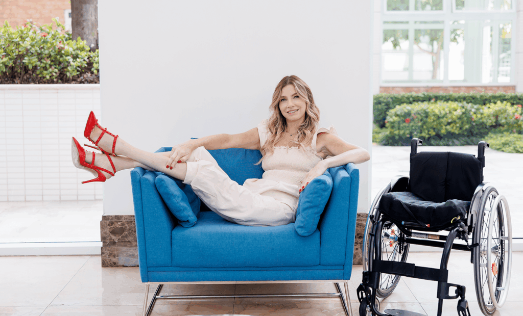 marketing. Foto na nossa embaixadora Andrea sentada em um poltrona com as pernas para o ar, ao lado da poltrona está sua cadeira de roda. Andrea veste um conjuto na cor bege e usa um salto alto na cor vermelha. 